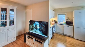 Uma sala de estar com televisão, uma cozinha com frigorífico e uma janela.