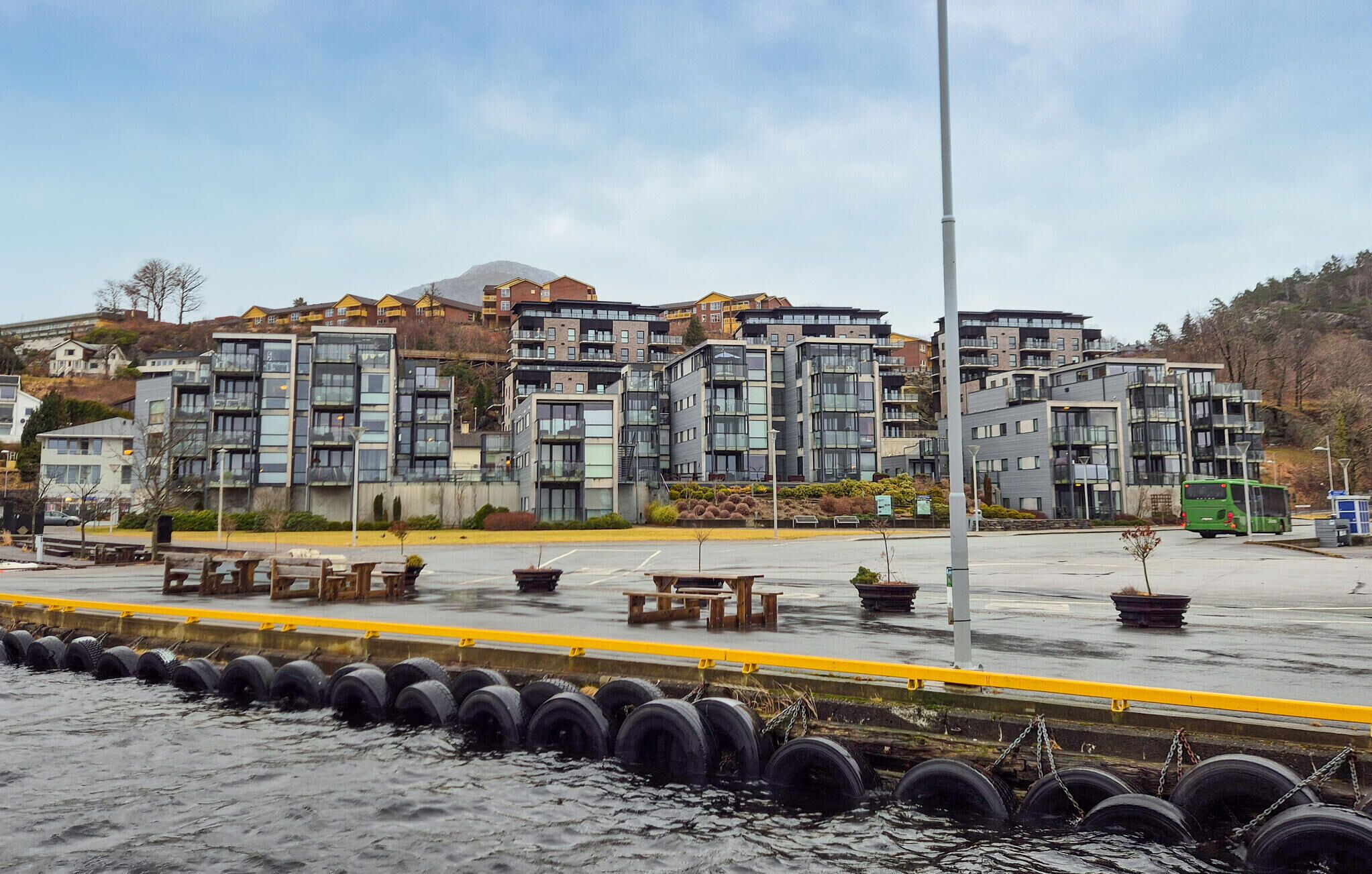 A waterfront residential area with modern apartment buildings, a pedestrian walkway, and a row of tires along the water's edge.