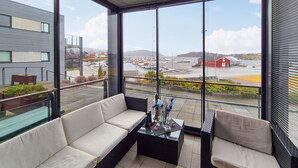 Uma varanda com sofá e mesa, grandes janelas com vista para um porto com barcos e edifícios.