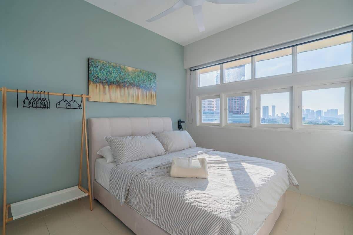 A bedroom with a bed, a painting, a window with a city view, and a ceiling fan.