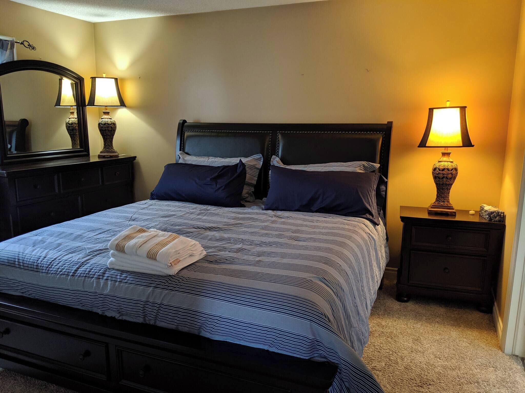 A neatly made bed with a patterned bedspread, two bedside lamps, a dark wooden headboard, and a small bedside table with a lamp.