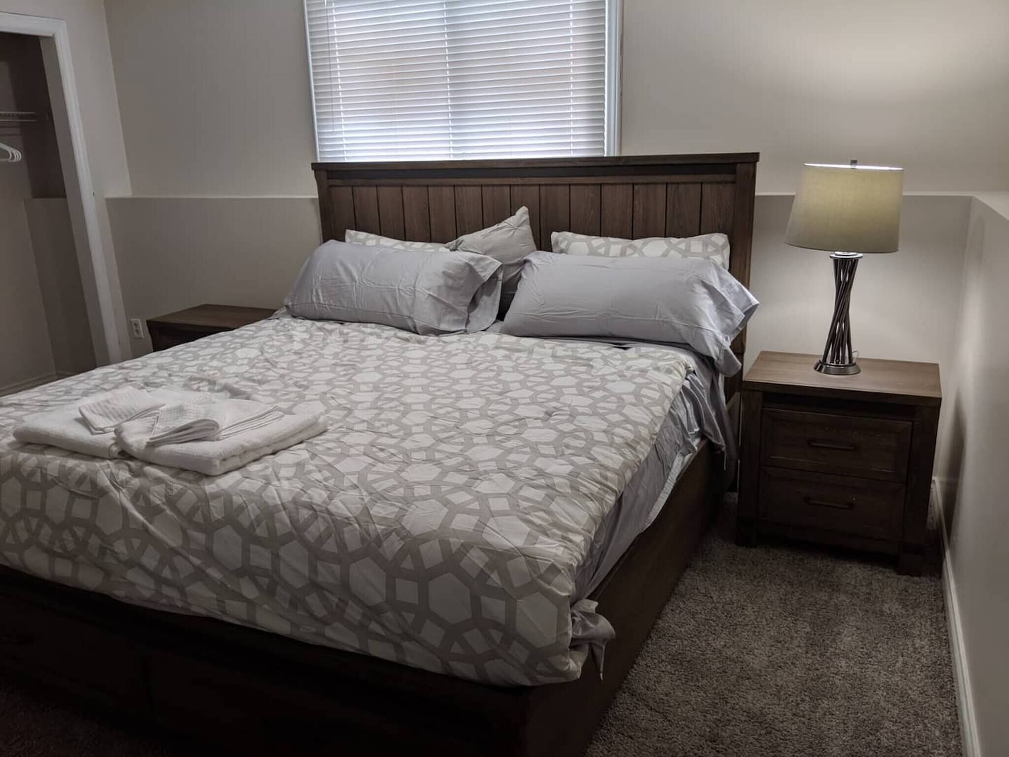 A neatly made bed with a patterned bedspread, two pillows, and a folded towel on the side. There is a nightstand with a lamp and a drawer.