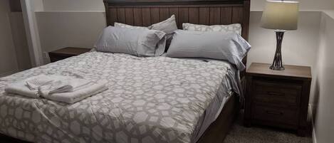 A neatly made bed with a patterned bedspread, two pillows, and a folded towel on the side. There is a nightstand with a lamp and a drawer.