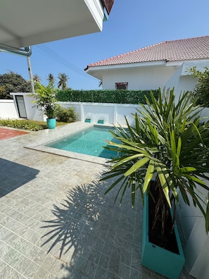 Une piscine dont le fond est recouvert de carreaux, entourée de plantes en pot et bordée d’un bâtiment blanc dont le toit est également recouvert de carreaux.