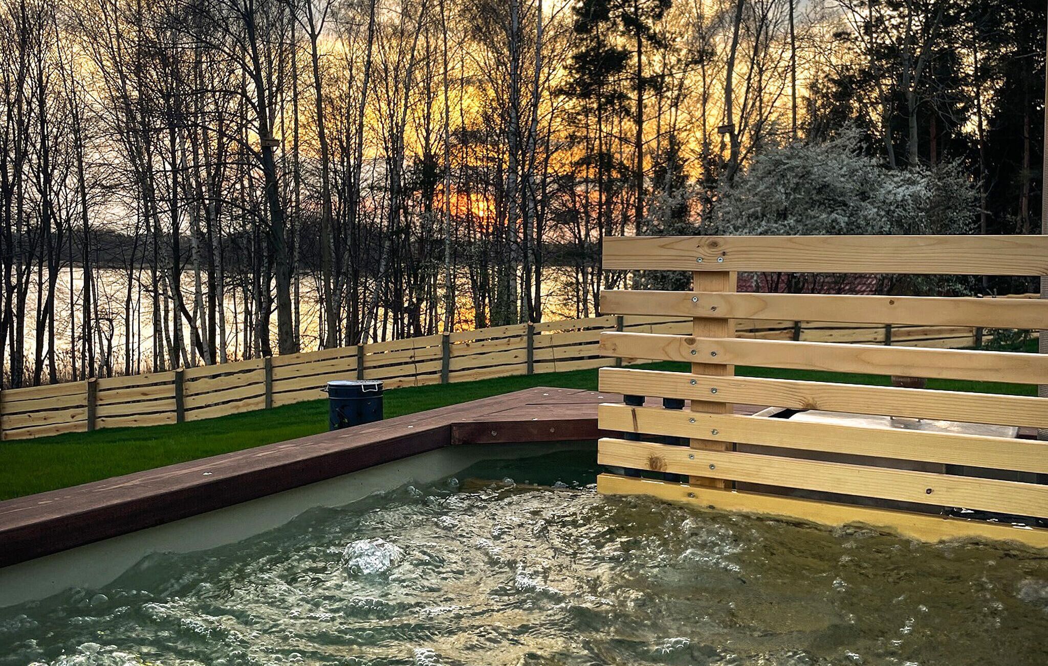 A hot tub area with a wooden deck, a grassy lawn, and a forested backdrop.
