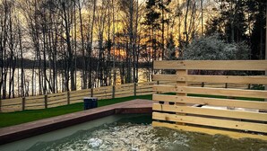 A hot tub area with a wooden deck, a grassy lawn, and a forested backdrop.