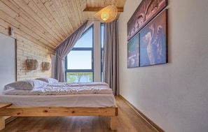 Un dormitorio con techo de madera, cama con ropa de cama blanca, un ventanal grande y un cuadro en la pared.