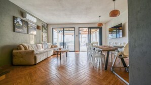 A spacious living area with a herringbone wooden floor, a sofa, a dining table with chairs, and a large window.