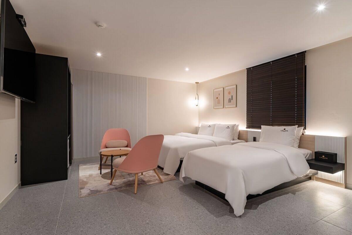 A modern hotel room with a bed, two bedside tables, a TV, a chair, and a window with blinds.