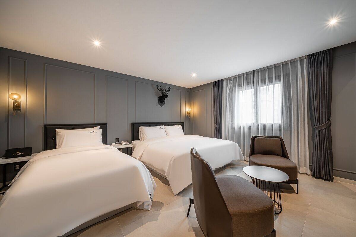 A hotel room with two beds, a sitting area with a chair and a small table, a mounted deer head on the wall, and a window with curtains.