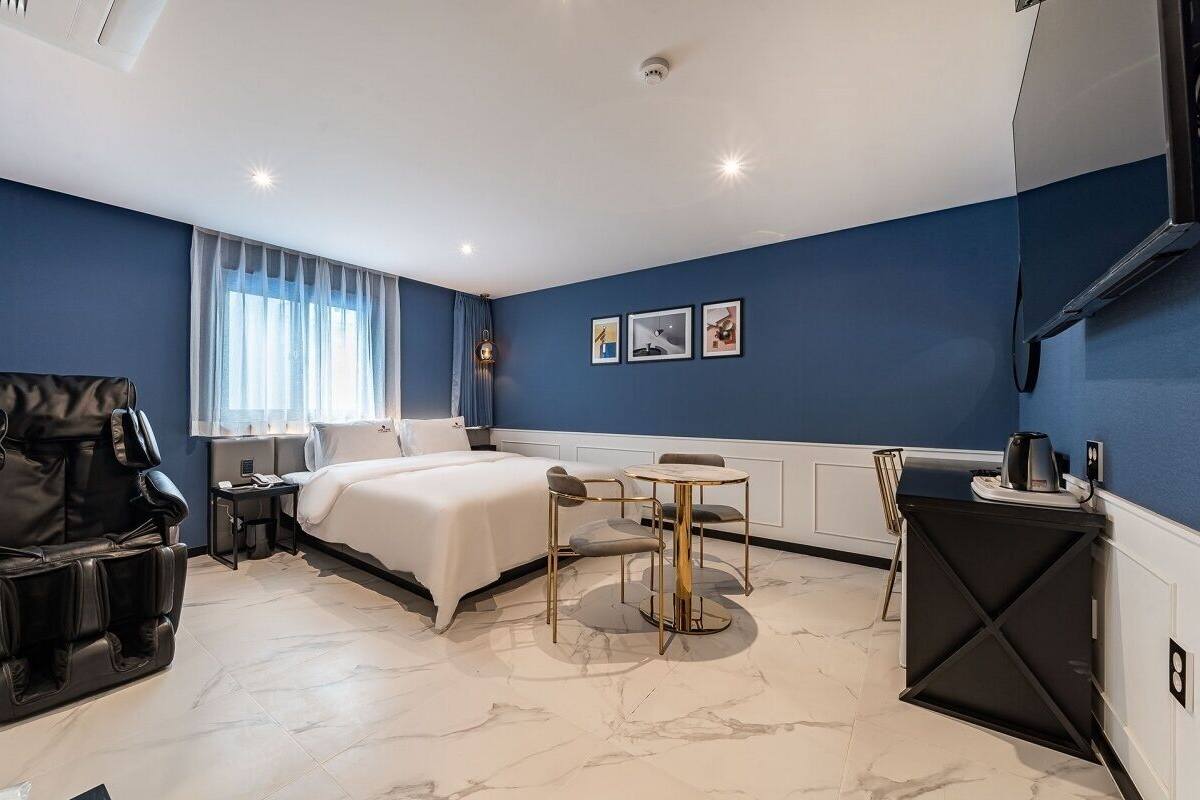 A modern hotel room with a bed, massage chair, small round table, and a kitchenette.