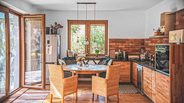 A modern kitchen with wooden cabinets, a dining table with wicker chairs, and a large window providing natural light.