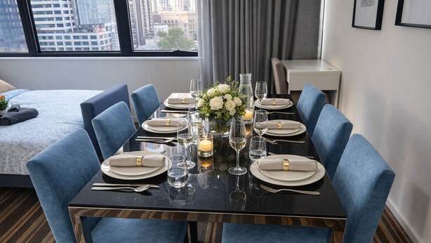 A dining table set for six with blue upholstered chairs, a vase of white flowers, candles, and clear glasses.