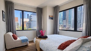 A hotel room with a bed, a sofa, a window with a city view, and a striped carpet.