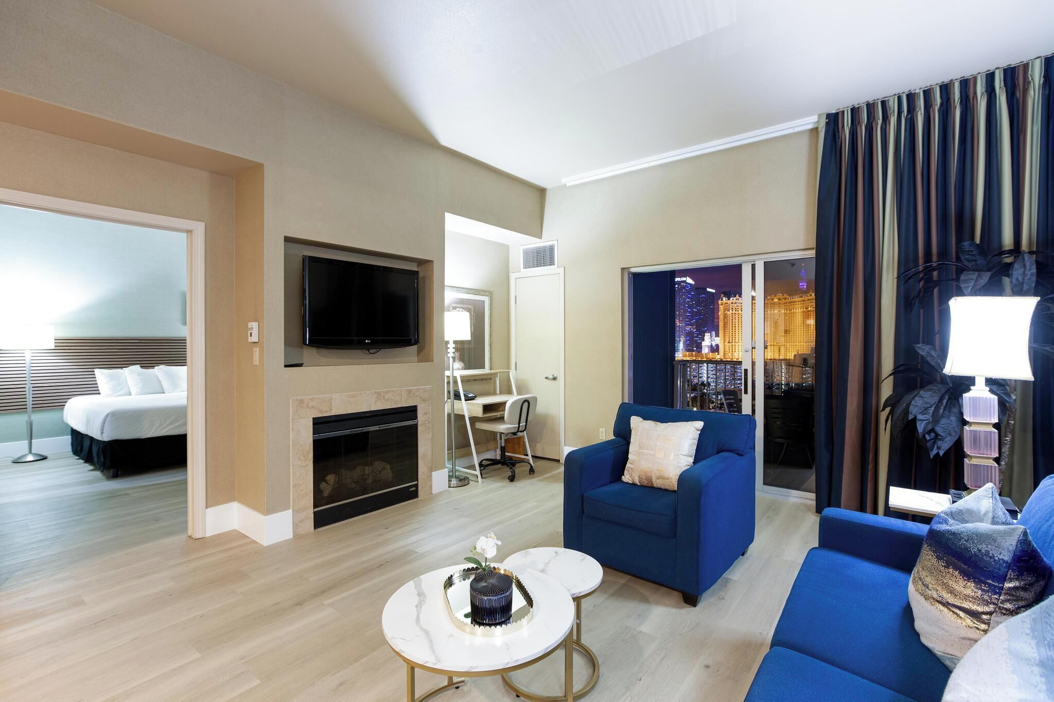 Habitación de hotel con cama, televisor, chimenea, sofá azul, mesa de centro y vista a un paisaje urbano a través de la ventana.