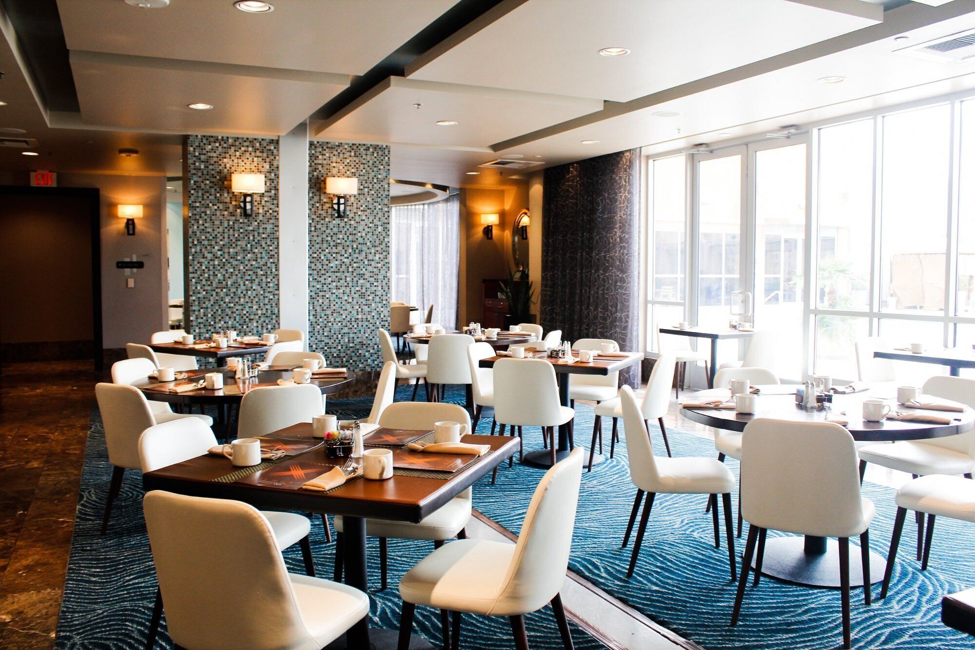 A hotel dining area with multiple tables and chairs, large windows, and decorative wall tiles.
