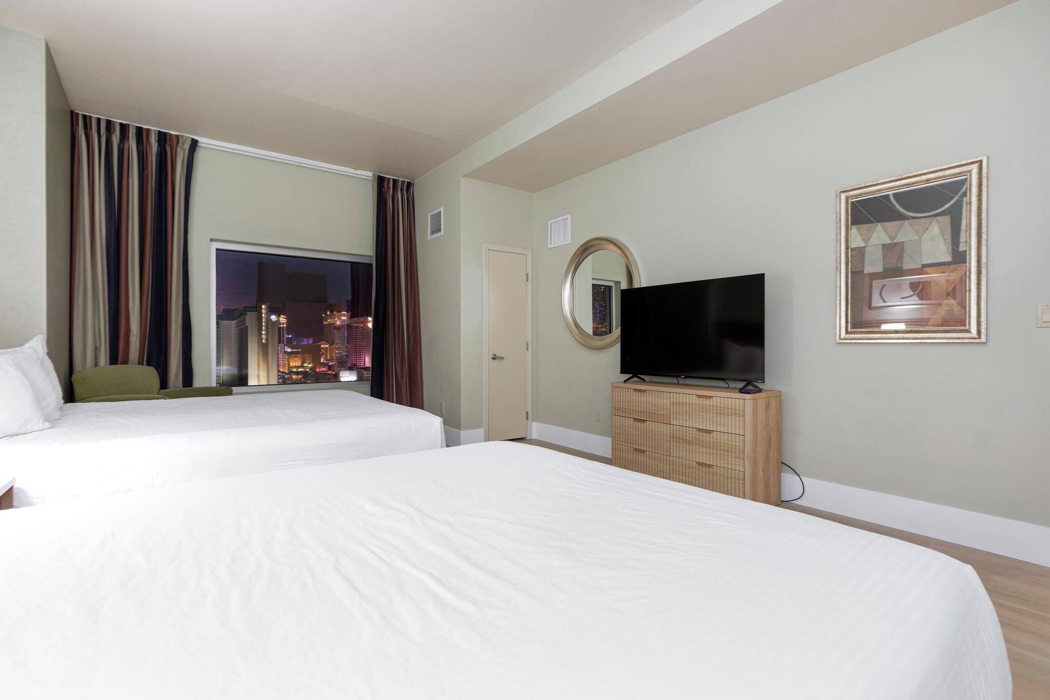 A hotel room with two beds, a television, a mirror, and a view of a cityscape at night.