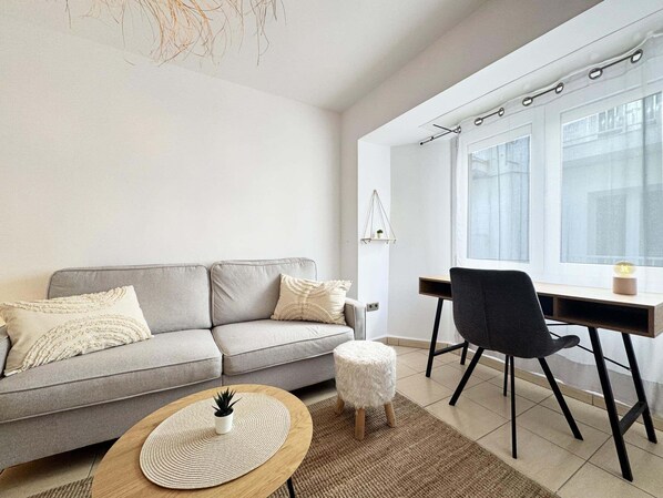 A modern living room with a grey sofa, a round coffee table with a plant, a small stool, and a desk with a chair.