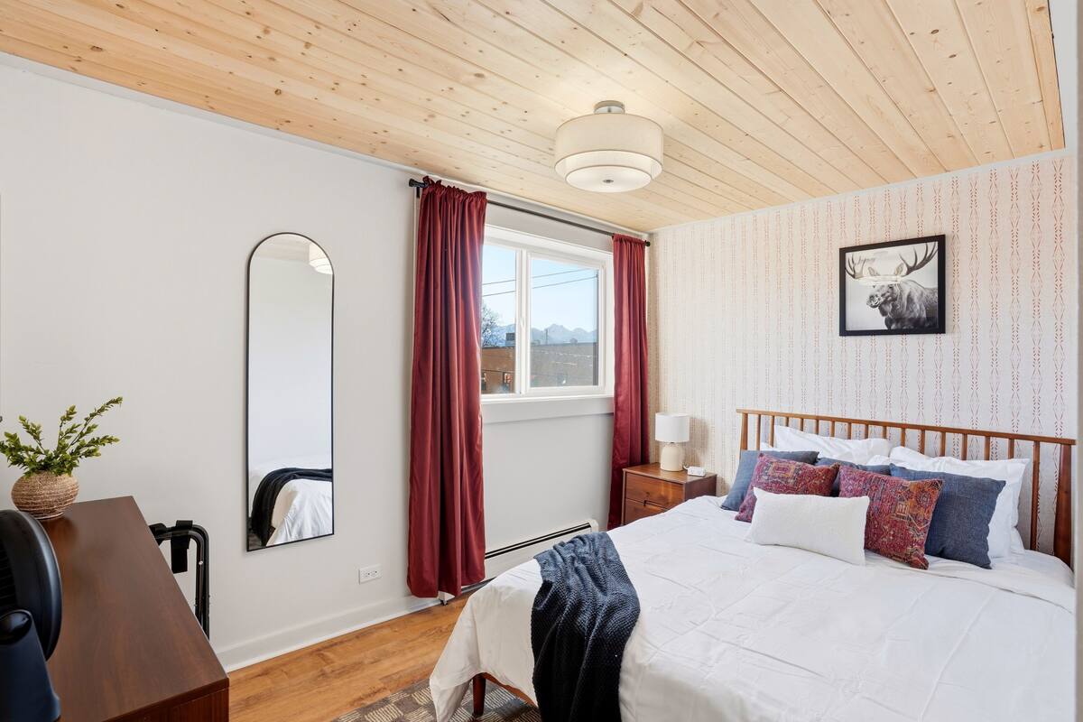 Ein Schlafzimmer mit Holzboden, einem Bett mit vielen Kissen, einem Nachttisch mit Lampe, einem Spiegel und einem fenster mit Vorhängen.
