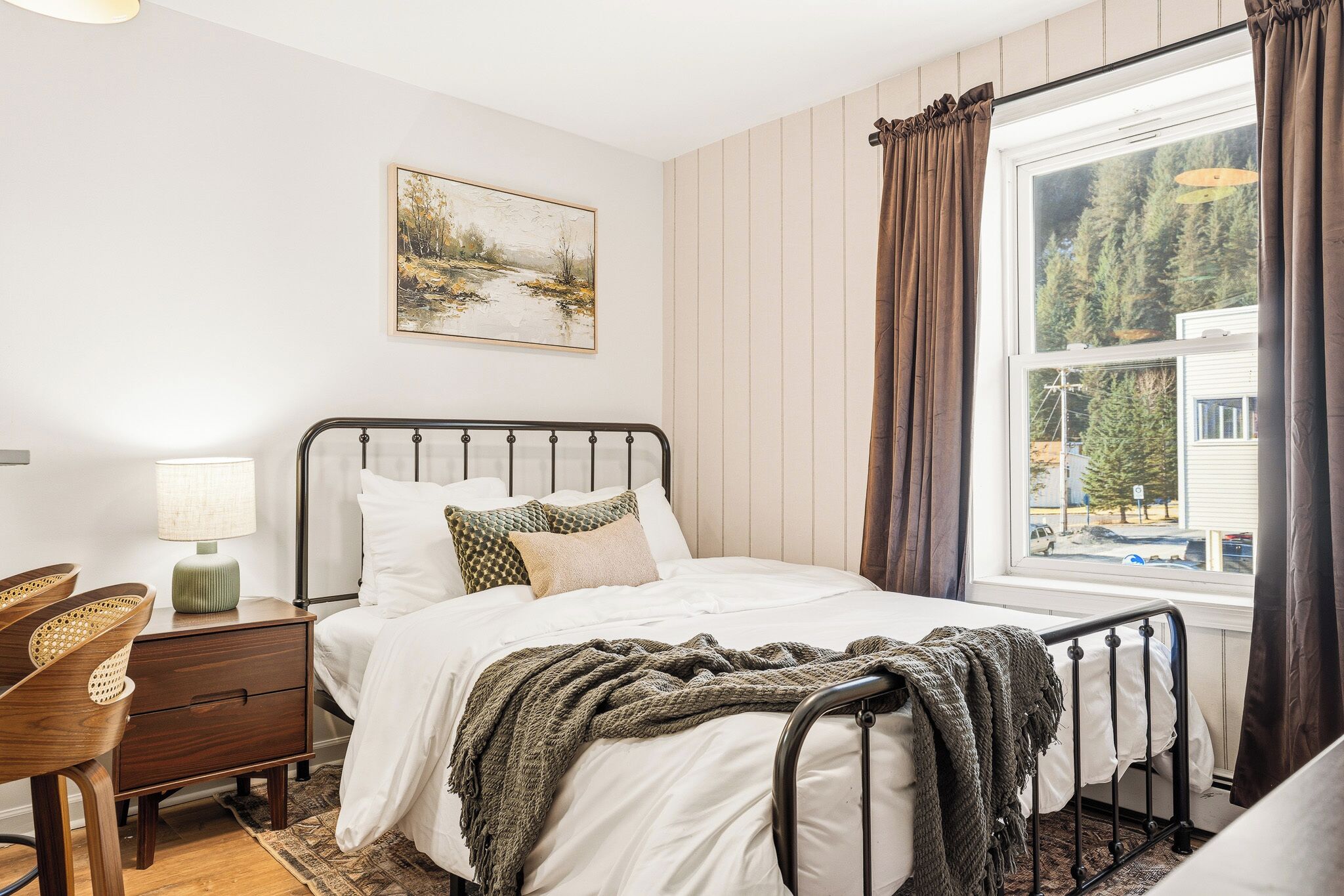 Quarto com cama, mesa de cabeceira, abajur e janela com cortinas.