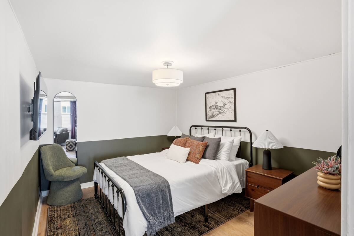 Ein Schlafzimmer mit Bett, Nachttischen, einem Sessel, einem Schreibtisch, einer Lampe, einem Spiegel und einem gerahmten Bild an der Wand.