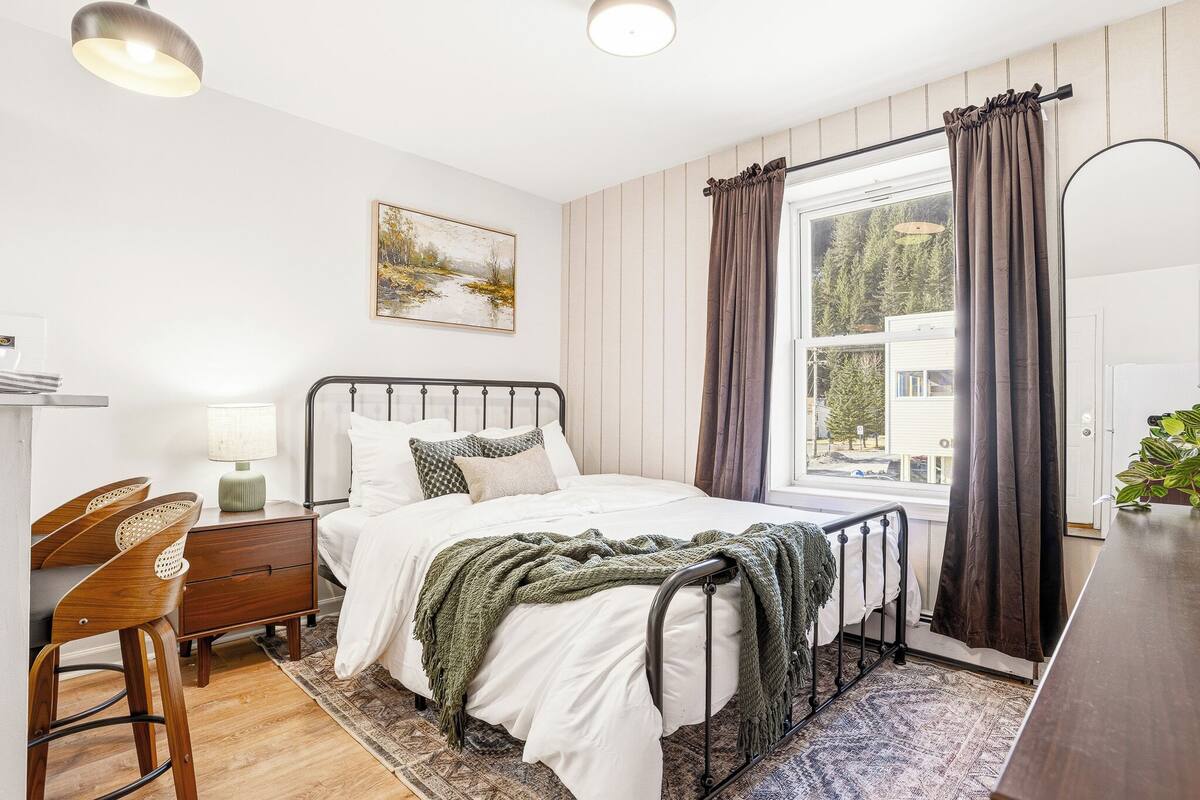 Ein Schlafzimmer mit Bett, Nachttisch, Lampe, Stuhl und einem Fenster mit Vorhängen.
