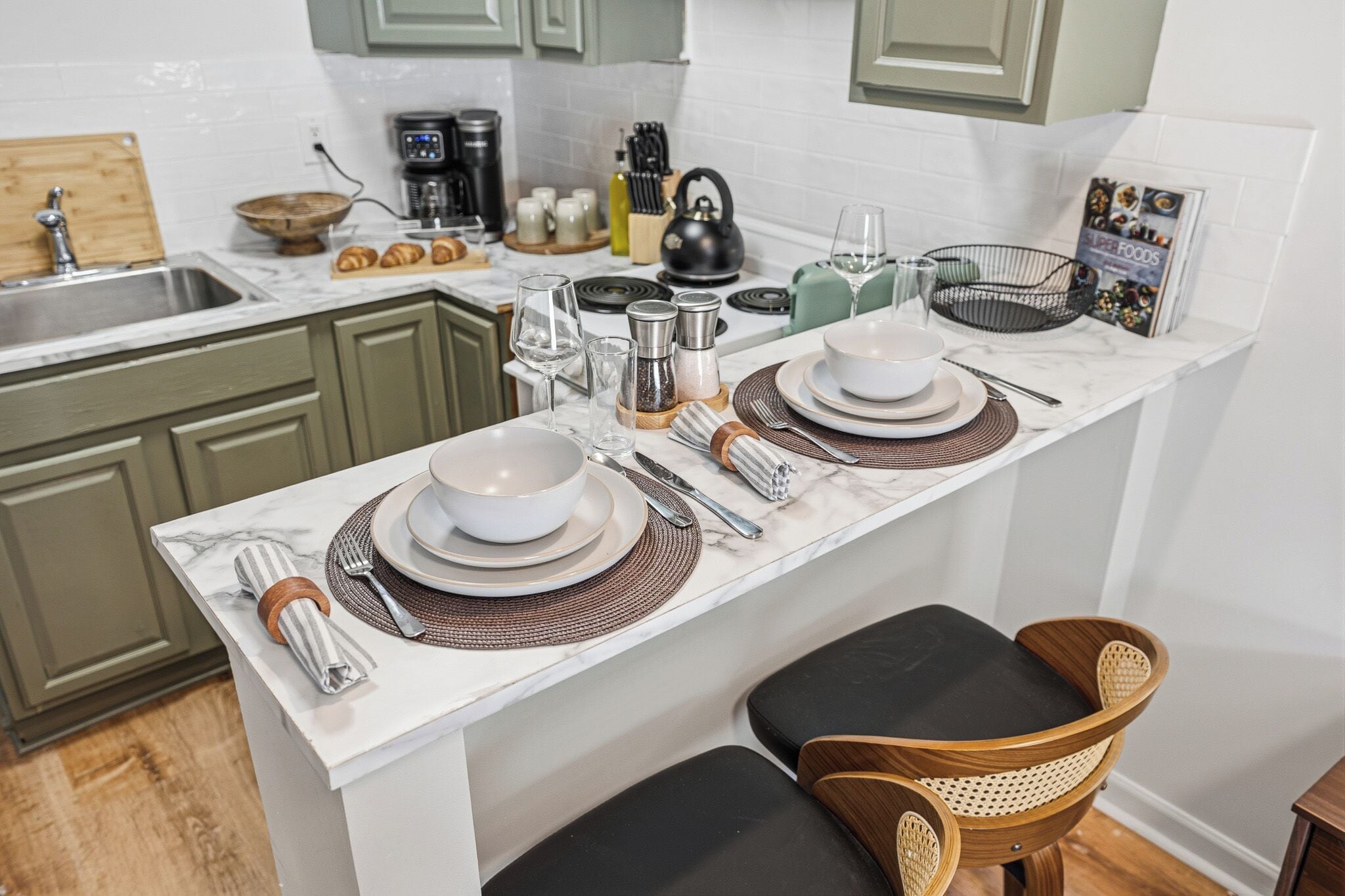 A kitchen with a marble countertop, green cabinets, a coffee maker, a kettle, and a dining area with a table set for two.