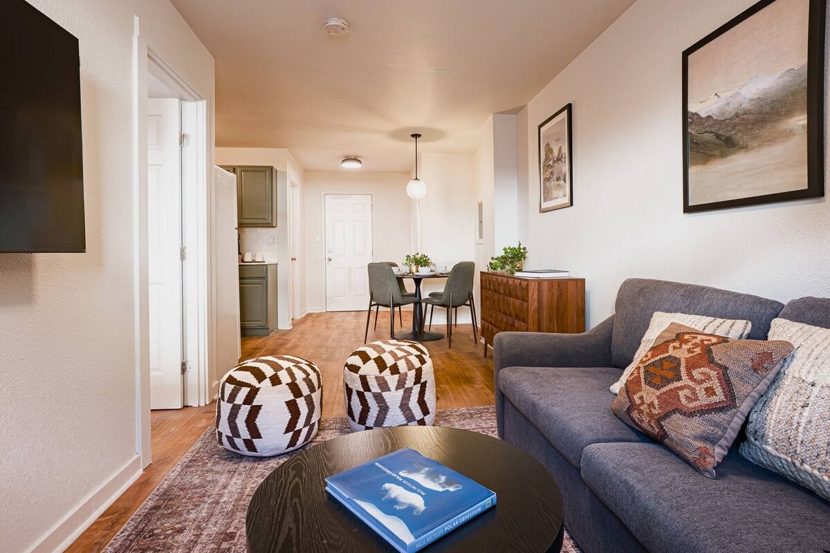 Ein modernes Wohnzimmer mit grauem Sofa, gemusterten Hocker, einem Couchtisch mit Buch, einem Essbereich mit Stühlen und einer Küche im Hintergrund.