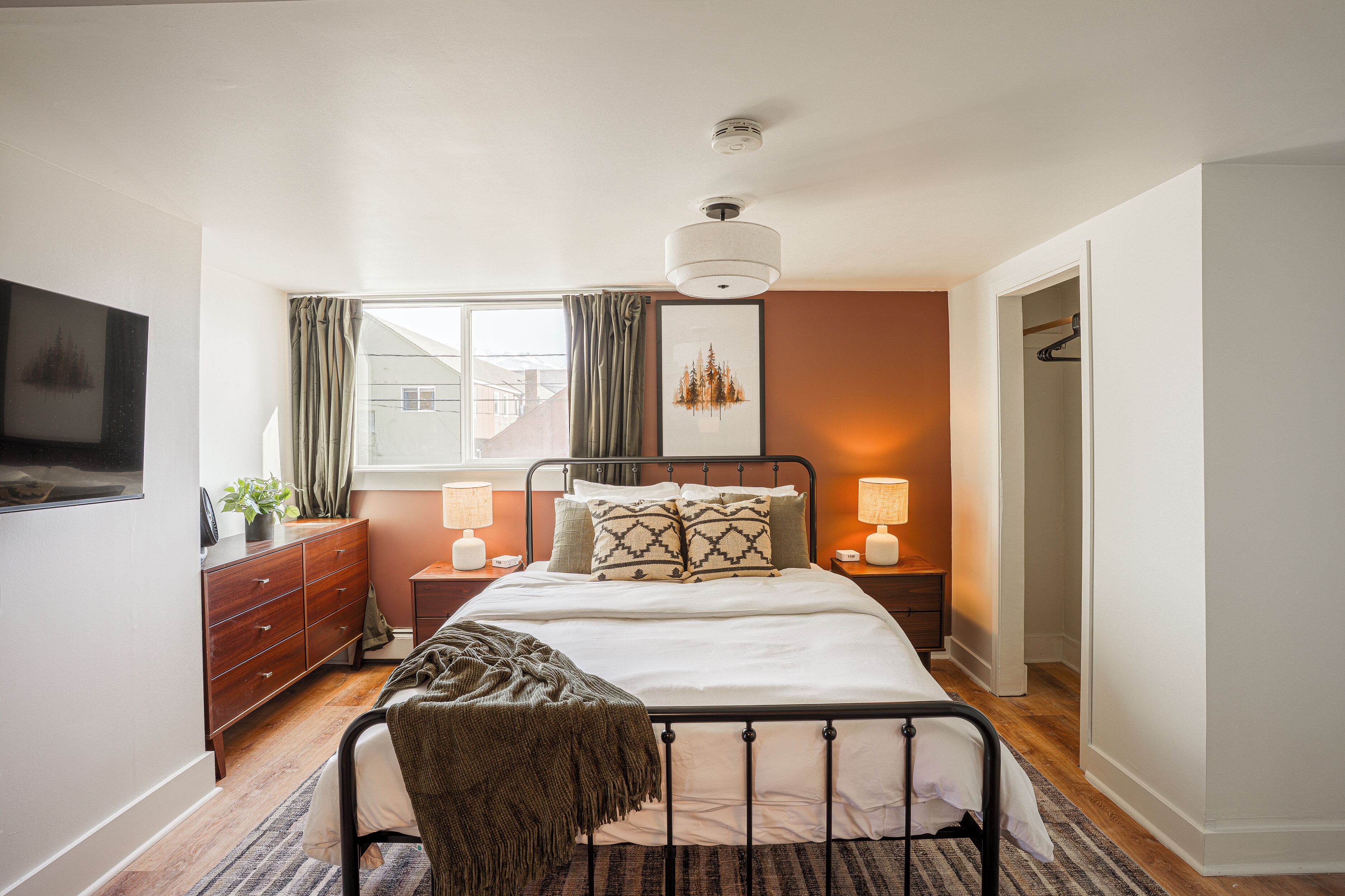A bedroom with a bed, bedside tables, a dresser, a window with curtains, a hanging light fixture, and a door leading to another room.