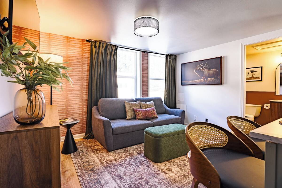 Ein modernes Wohnzimmer mit einer Couch, einem Hocker und Korbstühlen, einer Topfpflanze und einem Bild an der Wand.