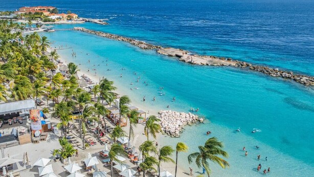 Un complejo turístico en la playa con un rompeolas rocoso, aguas turquesas cristalinas y palmeras.