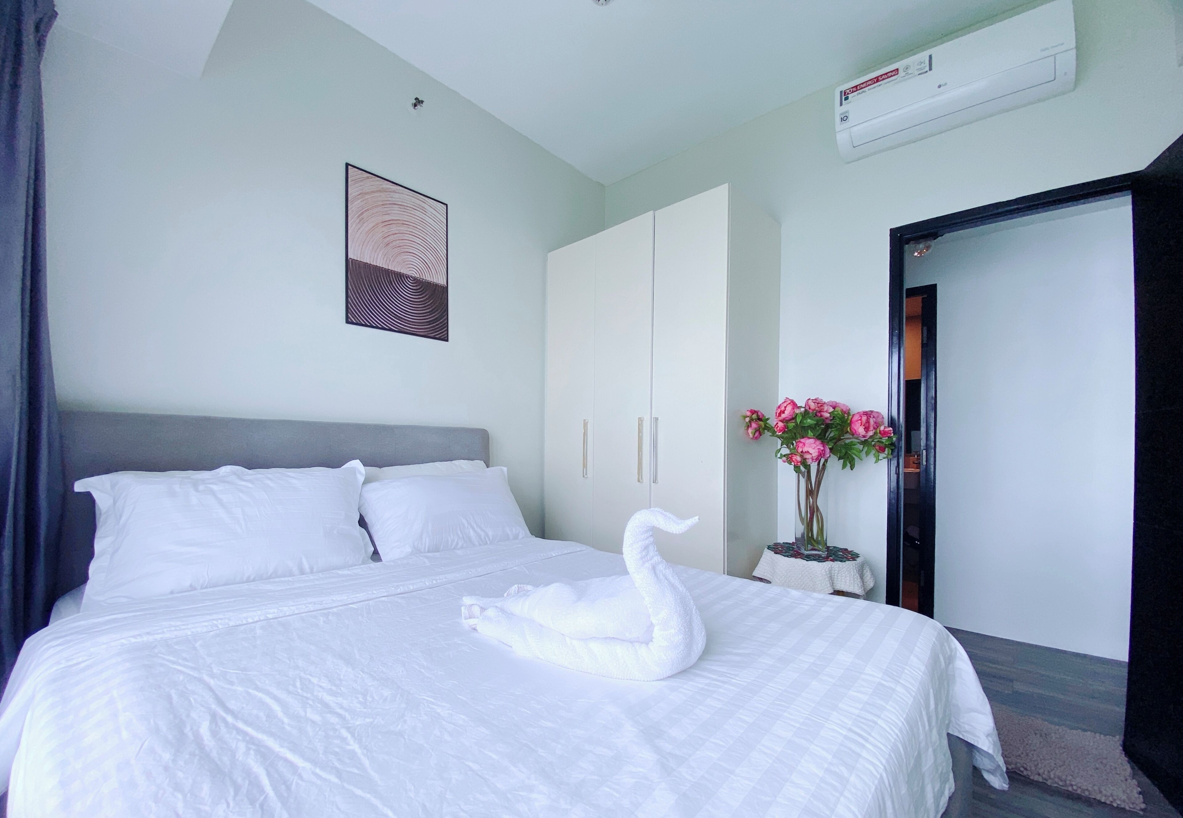 A hotel room with a bed, a white swan-shaped pillow, a wall-mounted air conditioner, a framed picture, and a vase with flowers.