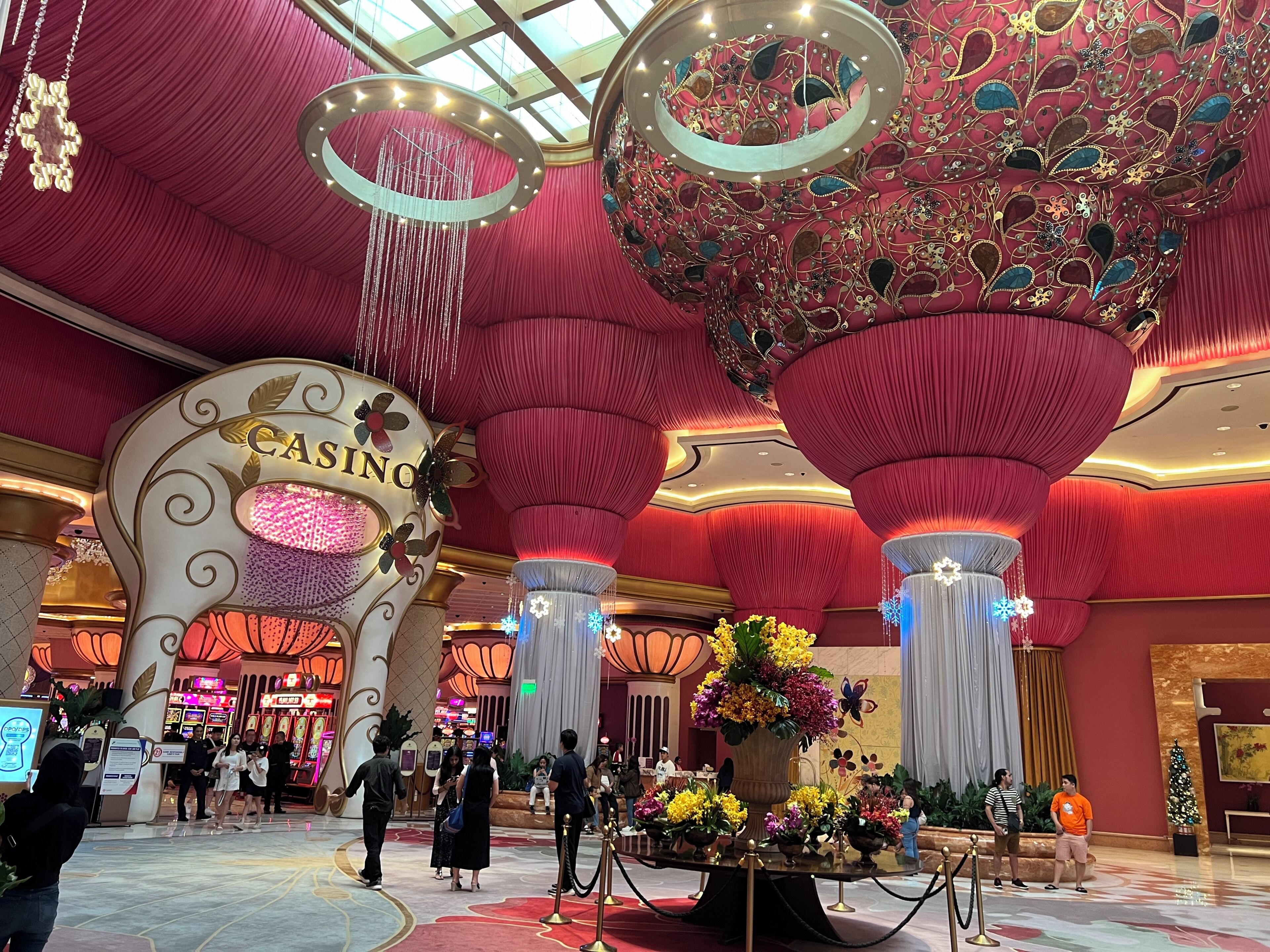 Interior de un casino con un llamativo letrero que dice 'CASINO', grandes candelabros decorativos y un arreglo floral central.