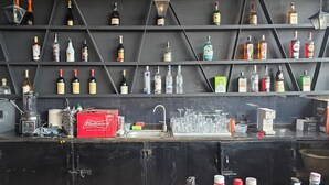 Zona de bar con estantes llenos de botellas de licor, una caja de Budweiser, vasos y un lavamanos.