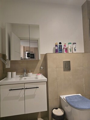 A modern bathroom with a white sink, mirror, and toilet. There are toiletries on the wall and a blue mat on the toilet. - Modern Studio in Accra near Airport & Mall (Accra)