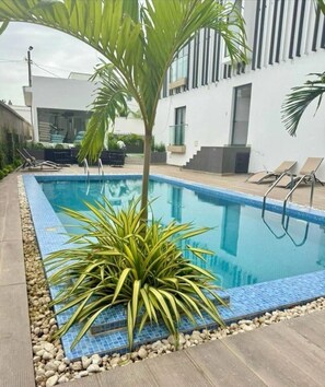 A swimming pool with a tiled edge, surrounded by a wooden deck and a small palm tree. - Modern Studio in Accra near Airport & Mall (Accra)