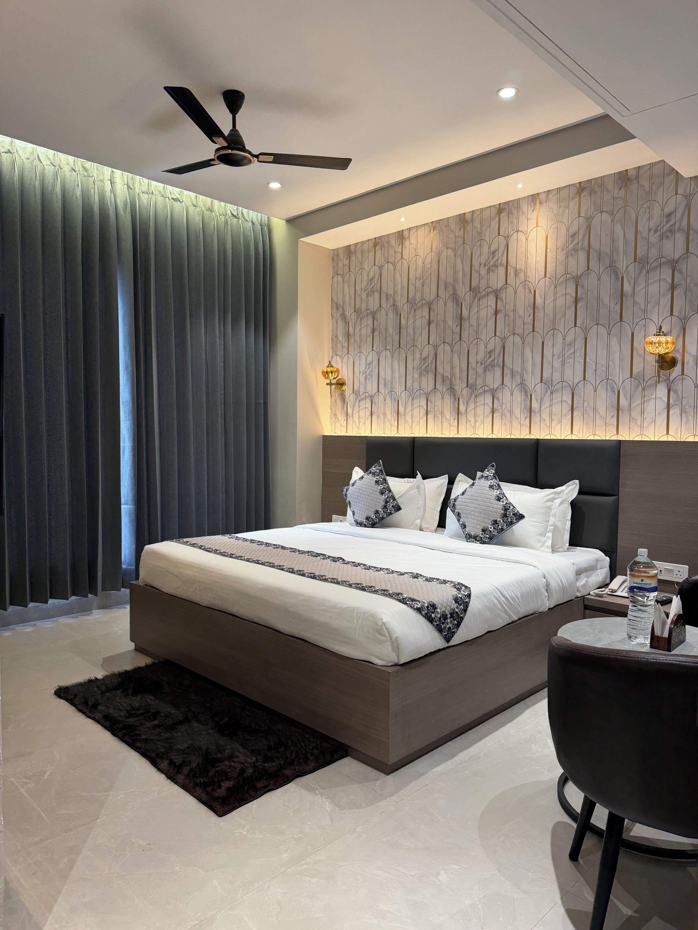 A modern bedroom with a large bed, a ceiling fan, a marble-patterned wall, and a small side table with a water bottle.