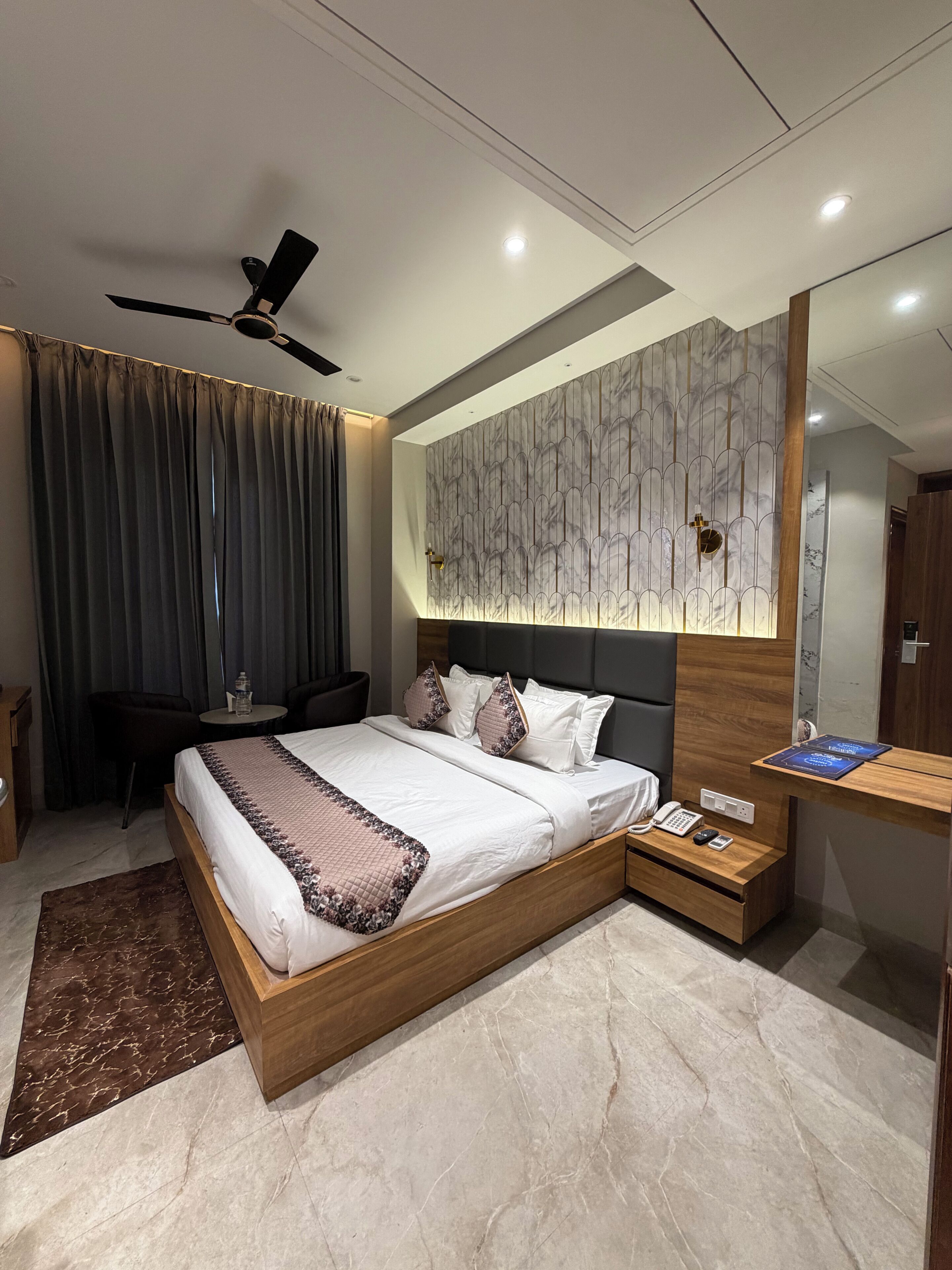 A modern hotel room with a large bed, a desk, a chair, a ceiling fan, and a mirror.