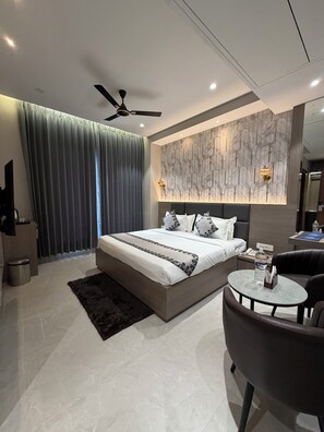 Une chambre moderne avec un grand lit, un ventilateur de plafond, un mur décoré d’un motif marbré, une petite table ronde et un fauteuil en cuir foncé.