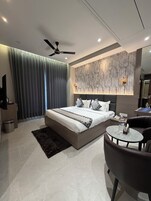 Un dormitorio moderno con una cama grande, un ventilador de techo, un muro con diseño de mármol, una mesita redonda y un sillón de cuero oscuro.