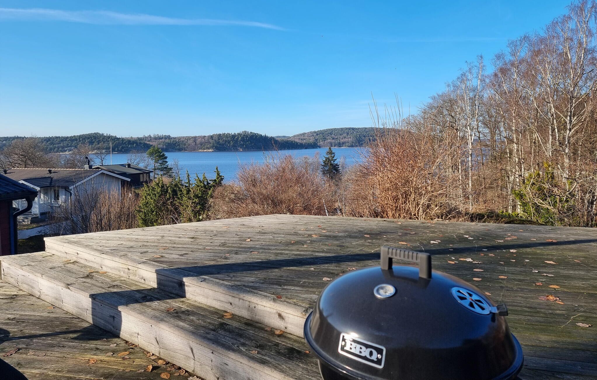 A BBQ grill on a wooden deck overlooking a lake with houses in the background.