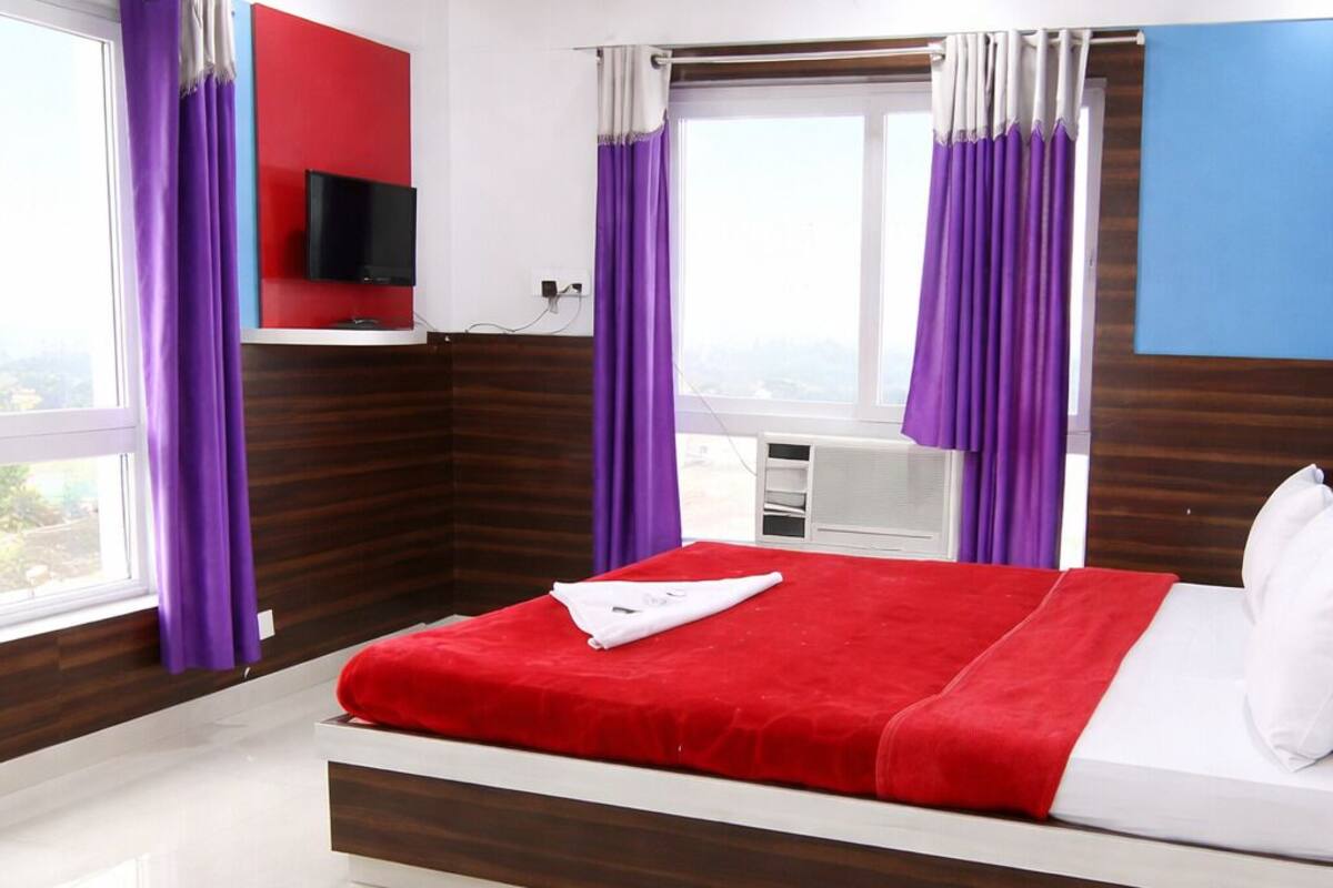 A hotel room with a bed, a TV mounted on the wall, and a window with purple curtains.
