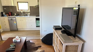 A living room with a wooden floor, a sofa, a TV, a table with remote controls, and a kitchen in the background. - Cosy Cottage at Anglers Reach close to Lake Eucumbene and Selwyn Snow Resort (Anglers Reach)