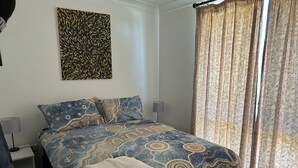 A bedroom with a patterned bedspread, a wall-mounted artwork, a bedside lamp, and a window with curtains. - Cosy Cottage at Anglers Reach close to Lake Eucumbene and Selwyn Snow Resort (Anglers Reach)