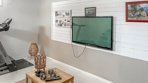 Ein Zimmer mit Laufband, Wandfernseher, kleinem Holztisch mit Fernbedienungen und Korkwand mit Fotos und Notizen.