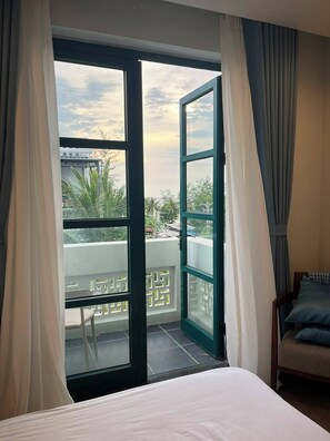 Habitación de hotel con cama, una silla y balcón con vista a palmeras y un edificio.