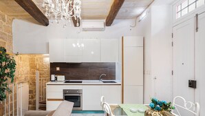 A modern kitchen with white cabinets, a built-in oven, and a stainless steel sink. There is a dining area with a green table and a vase of flowers. - Chic Comfy 1BR Maisonette Valletta Area (Floriana)