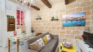 A cozy living room with a stone wall, a sofa, a dining table, and a television. - Chic Comfy 1BR Maisonette Valletta Area (Floriana)
