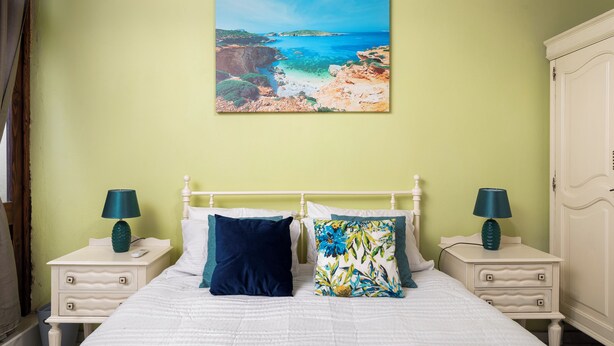 A bedroom with a bed, two nightstands, a wardrobe, and a painting of a coastal landscape. - Chic Comfy 1BR Maisonette Valletta Area (Floriana)