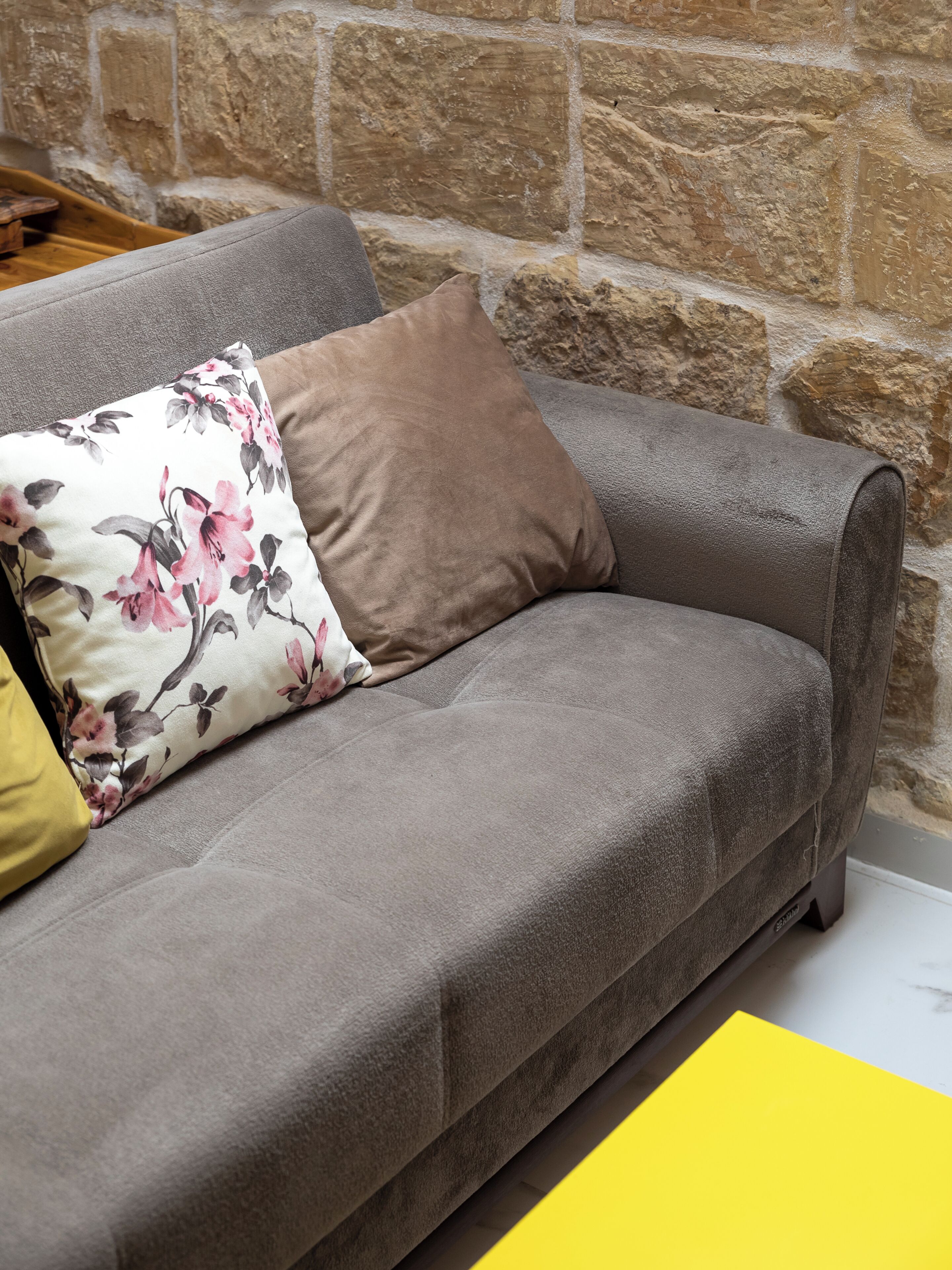 A grey sofa with a brown cushion and a floral pillow against a stone wall.